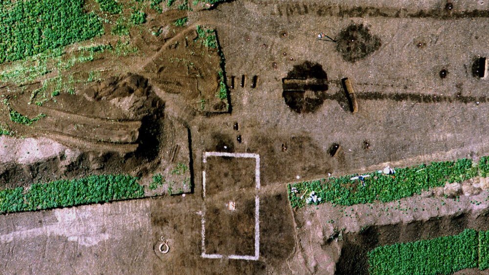 Abbildung 10: Deutlich erkennbar ist das rechteckige Betonfundament des Ziegelgebäudes mit dem mittigen Schornstein und dem nahe gelegenen Abwasserschacht. Dunkel heben sich verschiedene vorgeschichtliche Befunde vom anstehenden Boden ab. © Ingo Hoffmann.